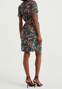 Woman wearing a knee-length, short-sleeve floral dress with black high-heeled sandals, standing against a plain background.