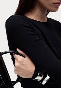 Schwarzes, geripptes Langarmoberteil, das mit einer schwarzen Handtasche getragen wird, die glatte Griffflächen und silberne Armreifen am Handgelenk aufweist.