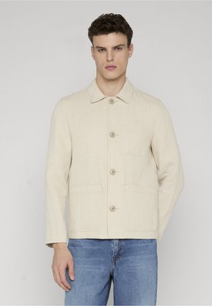 Mann trägt eine beigefarbene, gerippte Jacke mit Knöpfen und Kragen, kombiniert mit blauen Jeans, steht vor einem schlichten hellen Hintergrund.