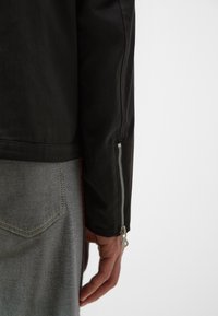 Zwarte leren jas met een gladde textuur, voorzien van een ritsmanchet en een minimalistisch ontwerp. Grijze denim eronder met contrasterende stiksels.