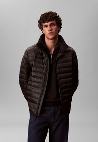 Must puffer-jakk lukuga ees, millel on kõrge krae ja tikitud disain, kombineeritud tumeda kampsuni ja teksapükstega.