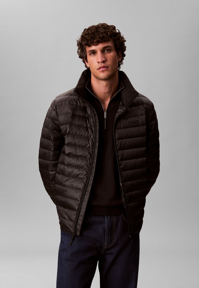 Must puffer-jakk lukuga ees, millel on kõrge krae ja tikitud disain, kombineeritud tumeda kampsuni ja teksapükstega.