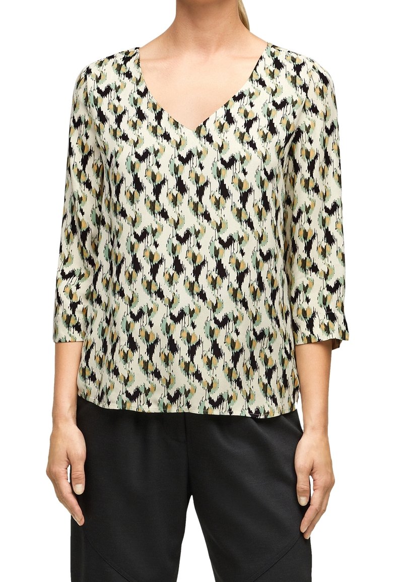 Femme portant un chemisier en V avec un motif abstrait multicolore et des manches trois-quarts, associée à un pantalon noir.
