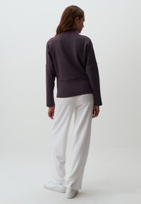 Maglione viola con collo alto, spalle scese e vestibilità rilassata, abbinato a pantaloni bianchi a gamba larga e sneakers bianche.