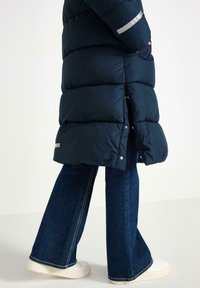 Cappotto puffer blu con design trapuntato, dettaglio riflettente sulla manica, abbinato a jeans in denim scuro a zampa d'elefante e sneakers bianche.