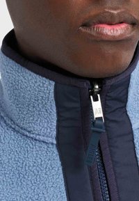 Svijetloplava fleec jakna na zip s tamnoplavim detaljima. Ima visoki ovratnik i teksturiranu tkaninu, a zupčanik za zatvaranje označen je sa "ANGLE 1853."