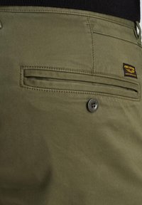 Pantalons vert olive avec une poche arrière à fermeture par bouton et un tissu texturé. Étiquette cousue près de la poche.