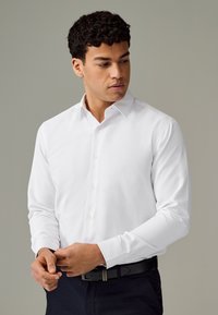 White long-sleeve shirt with a subtle textured pattern, button-down collar, and buttoned cuffs, paired with a dark belt and pants.