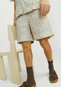 Pantalones cortos texturizados en color beige claro con un patrón de hojas, combinados con calcetas de rayas marrones oscuras y sandalias marrones. Silla minimalista beige en el fondo.