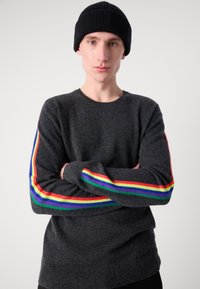 Tummanharmaa neulepusero, jossa on ribbitekstuuri. Hihassa on värikkäitä raitoja punaisena, keltaisena, vihreänä, sinisenä ja purppurana. Musta neulottu pipo.