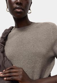 Jasnobrązowy prążkowany sweter z okrągłym dekoltem, zestawiony z ciemnobrązowym pikowanym akcentem na ramionach. Widoczne złote kolczyki koła.