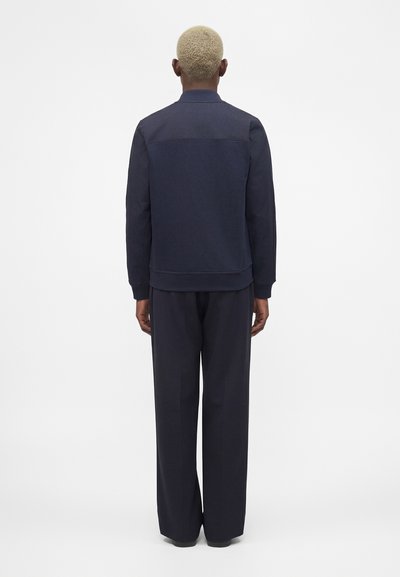 Άτομο με κοντά ξανθά μαλλιά στέκεται πλάτη προς την κάμερα, φορώντας σκούρο μπλε σακάκι και αντίστοιχο άνετο παντελόνι σε μονόχρωμο φόντο.