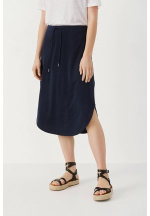 Frau trägt einen marineblauen Midirock mit Kordelzug und seitlichem Schlitz sowie schwarze, Riemchen-Espadrilles vor einem einfarbigen, hellen Hintergrund.