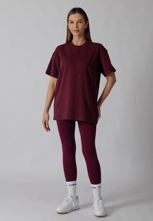 Kvinde stående iført en løs mørkerød t-shirt, matchende leggings, hvide sokker med sort tekst og hvide sneakers mod en ensartet grå baggrund.