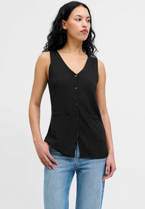 Femme aux longs cheveux noirs portant un gilet noir sans manches boutonné et un jean bleu clair, debout devant un fond blanc uni.