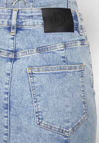 Jean en denim bleu clair avec une poche arrière cousue et une étiquette de marque en cuir noir au-dessus de la poche sur la ceinture.