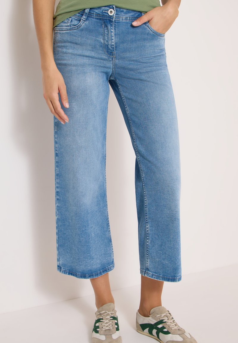 Femme portant un jean bleu clair court à coupe large, les mains dans les poches, et des baskets blanches avec des touches de vert.