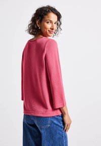 Rosa Fleece-Oberteil mit Dreiviertel-Ärmeln, runder Ausschnitt und strukturiertem Stoff. Kombiniert mit blauen Jeans, die eine lässige Passform und einen entspannten Stil zeigen.