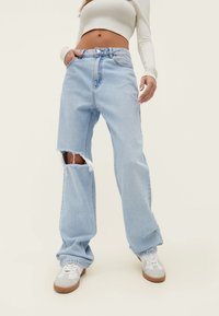 Vaqueros de denim desgastado de color azul claro con corte holgado. Presentan un agujero deshilachado en la rodilla izquierda y una cintura con botones. Llevados con zapatillas blancas.