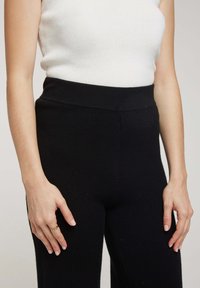 Pantalons en tricot noir avec une taille haute, texture douce et coupe ajustée. Le modèle porte un débardeur à côtes blanc et une bague en or à la main droite.