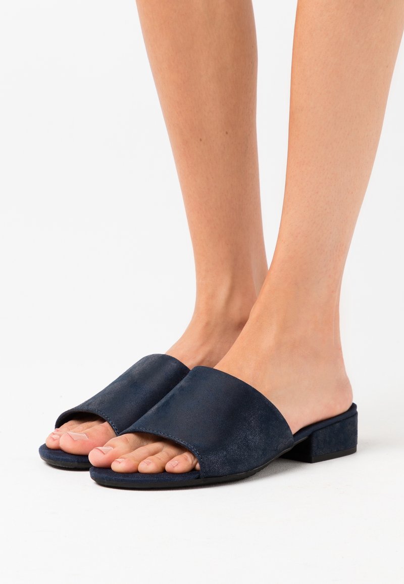 Pieds portant des sandales à talons blocs bas ouvertes à l'avant, de couleur bleu marine, sur une surface blanche.