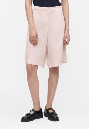 Kvinde iført knælange, lyserøde shorts med blonder i siderne, sorte Mary Jane-sko og en sort top, stående på en hvid baggrund.