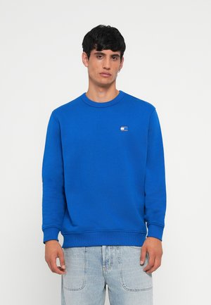 Blauwe katoenen sweatshirt met een ronde hals, geribbelde manchetten en tailleband; bevat een klein merkn logo aan de linkerkant van de borst. Gedragen met lichtblauwe jeans.