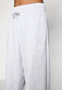 Heledad hallid dressipüksid pehmest materjalist, millel on elastne vöökoht ja nöör, ning diskreetne valge Adidas'i logo küljel.