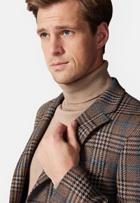 Brązowa marynarka w kratę z niebieskimi akcentami, teksturowany materiał i beżowy golf pod spód. Zbliżenie na górną część ciała mężczyzny, ukazujące detale.