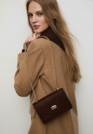 Borsa a tracolla in suede marrone con catena dorata, forma rettangolare e chiusura a patta, indossata sulla spalla su uno sfondo di un cappotto beige.