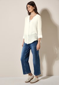 Hvid, struktureret langærmet skjorte med V-hals og buet kant, parret med brede blå denimbukser og beige sneakers.
