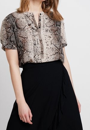 Femme portant un chemisier manches courtes à motif peau de serpent, rentré dans une jupe portefeuille noire taille haute avec un détail volanté.