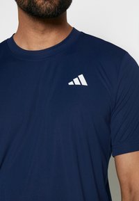 Marineblå atletisk t-shirt i mjukt tyg, med en vit triangulär logotyp på övre vänstra sidan och rund halsringning.