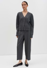 Femme se tenant devant un fond uni, portant un cardigan boutonné gris foncé, un pantalon gris ample et des ballerines noires à bout pointu.