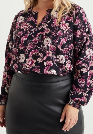 Kobieta w czarnej bluzce w kwiaty z długim rękawem, z różowymi i białymi kwiatami, włożonej do czarnej, wysokiej spódnicy z skóry.