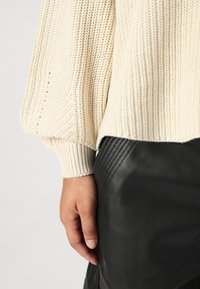 Beige gestrickter Pullover mit gerippten Bündchen, kombiniert mit schwarzen Lederhosen mit vertikalen genähten Akzenten. Hand auf dem Taillenbund.