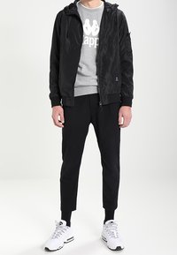 Sort hættejakke med en glat finish, grå sweatshirt med logo, sorte tilspidsede bukser og hvide sportssko med sorte detaljer.