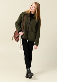 Giacca in pile verde oliva con colletto, bottoni neri, due tasche frontali, abbinata a jeans neri e stivali alla caviglia neri.
