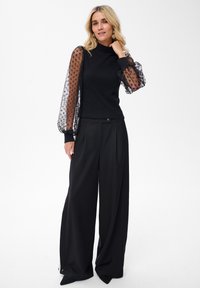 Czarny sweter z prześwitującymi, kropkowanymi balonowymi rękawami, w połączeniu z czarnymi spodniami o szerokich nogawkach i wysokim stanem, z delikatnym wykrojem z przodu.