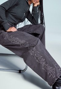 Šedé široké džínsy s mäkkou, textúrovanou tkaninou. Obsahujú dekoratívny perlový vzor pozdĺž bočných švov. Skombinované s tmavým zipsovým topom.