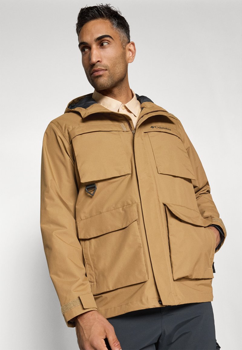 Chaqueta impermeable color marrón claro con capucha, grandes bolsillos frontales y puños ajustables. Presenta un forro interior negro en contraste y un logo.