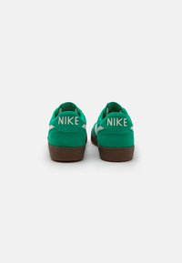 Zaļi zamša sporta apavi ar brūnu gumijas zoli, uz papēža ir Nike zīmols, un tiem ir elegants dizains ar izliekumiem.