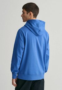 Felpa blu in cotone con cappuccio regolabile, polsini e orlo a coste, caratterizzata da una texture liscia e una vestibilità comoda, indossata con pantaloni scuri.