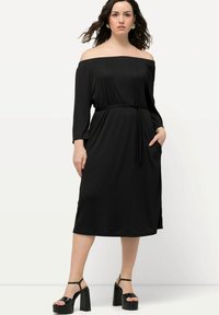 Czarna sukienka bez ramiączek z długimi rękawami, wiązanym w talii, z kieszeniami i zwiewną sylwetką. Modelka ma na sobie czarne sandały na platformie.