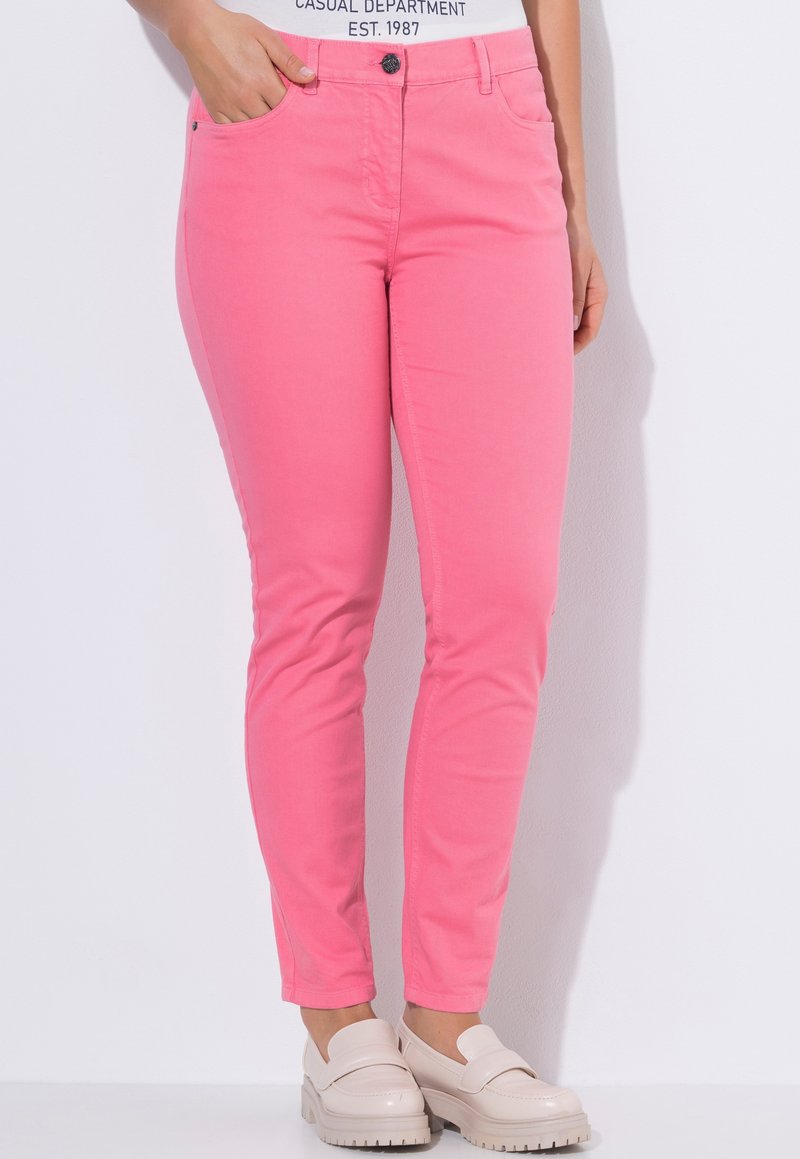 Close-up af en person iført tætsiddende pink bukser, hvide afslappede sko og en hvid skjorte med trykt tekst, stående mod en hvid væg.