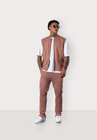 Hellrosa ärmellose Jacke über einem weißen T-Shirt, kombiniert mit hellrosanen Cargo-Hosen und weißen Sneakern. Sonnenbrille getragen, Tattoos sichtbar.