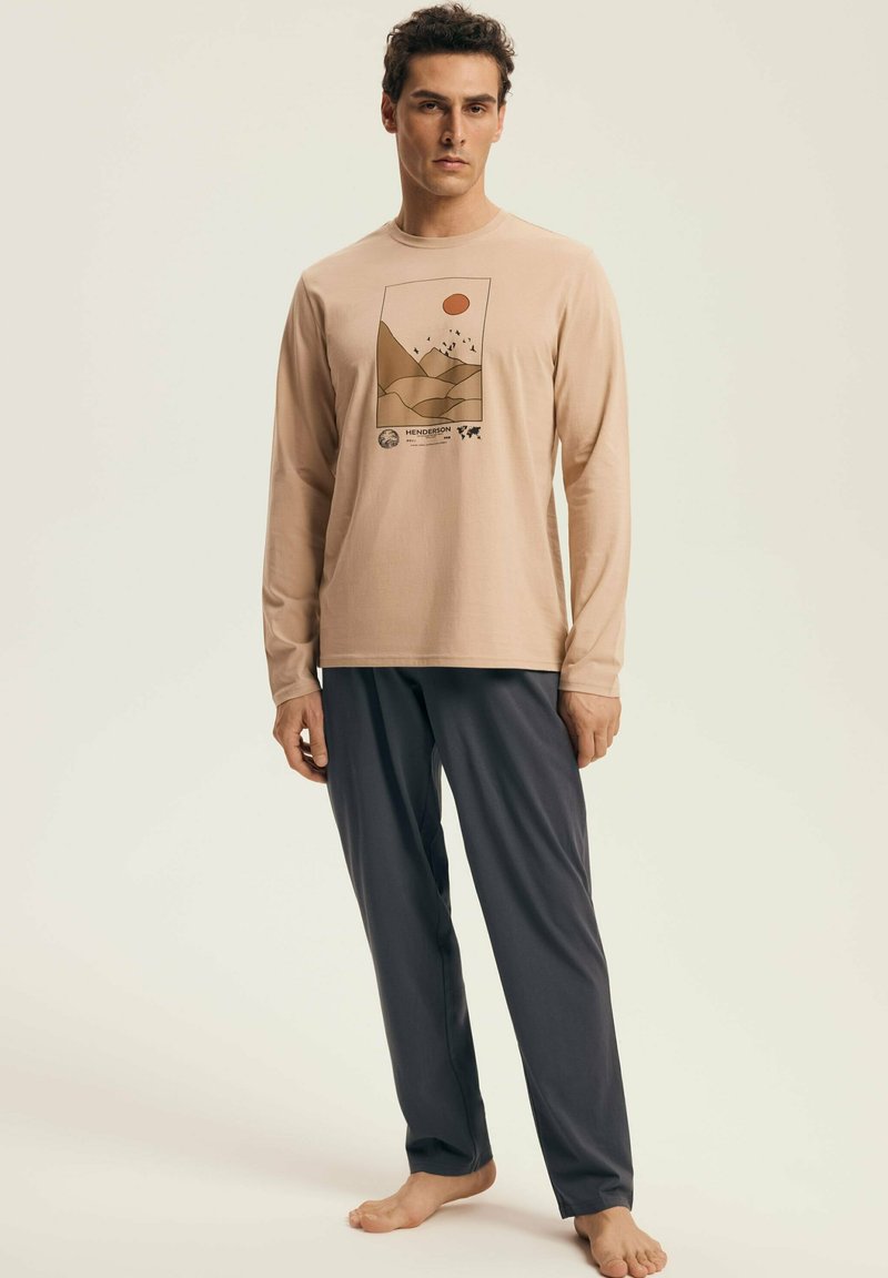 Young man standing barefoot wearing beige long-sleeve shirt with mountain print and dark gray pants against plain background.