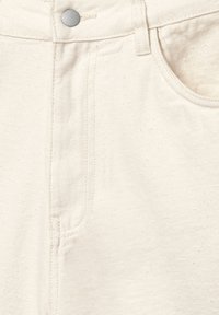 Denim di cotone beige con una finitura testurizzata, dotato di una tasca frontale, chiusura con bottone metallico e dettagli di cuciture a contrasto.