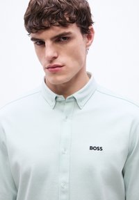 Νέος άνδρας με κοντά σγουρά μαλλιά, φοράει γαλάζιο πουκάμισο με κουμπιά και κέντημα "BOSS" στο στήθος, πάνω σε λευκό φόντο.