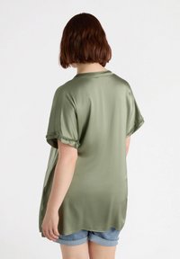 Blusa verde oliva a maniche corte con una texture setosa, caratterizzata da maniche volant e una vestibilità rilassata. Orlo curvo e spacco laterale sono visibili.